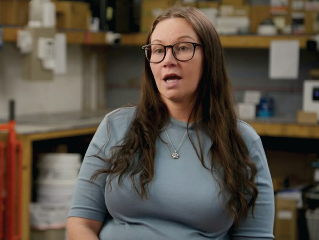 White woman with glasses and blue shirt discussing her experience.