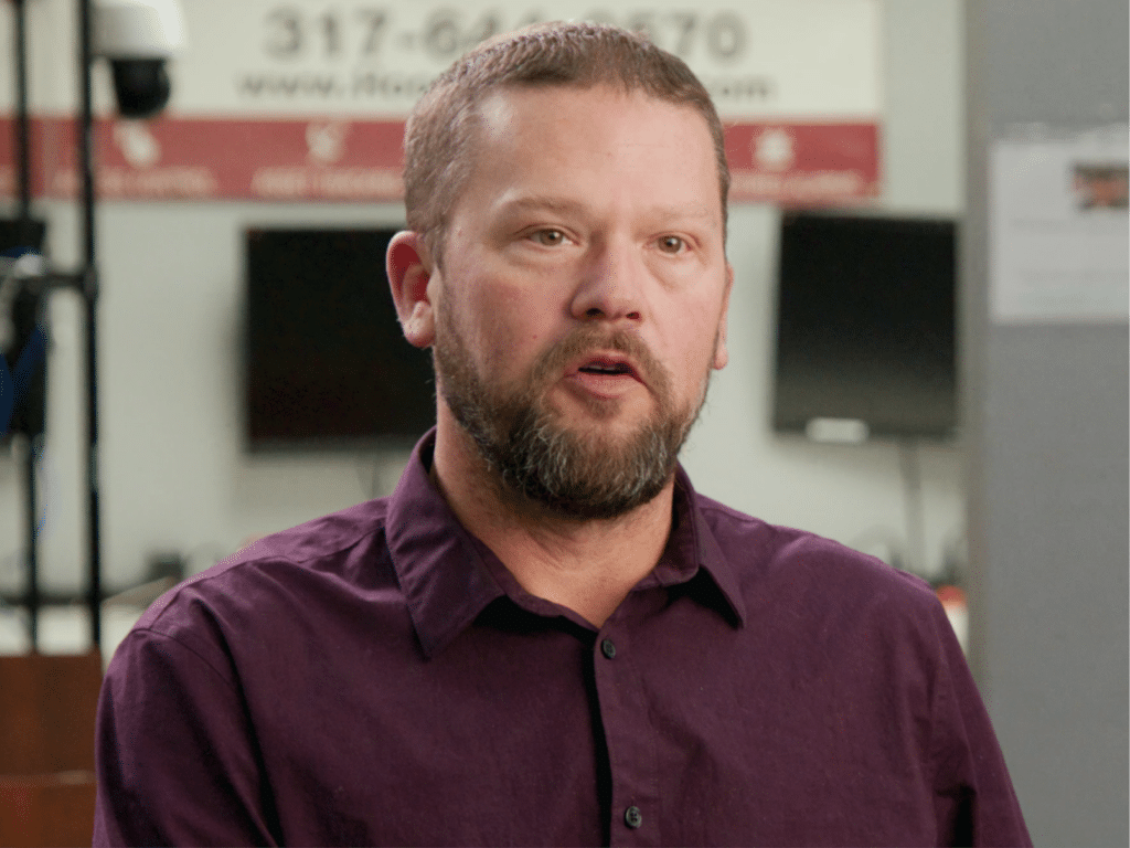 A man in a deep purple shirt talking about his experience